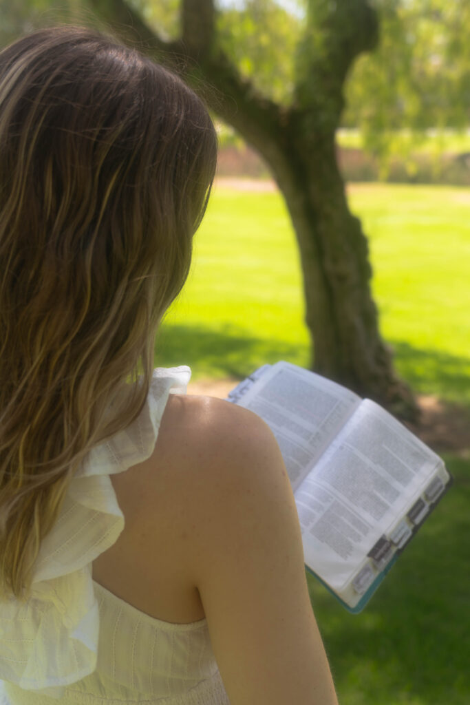 looking over the shoulder of a woman reflecting under the shade of a tree reading her bible, symbolizing discernment and personal faith after leaving a controlling Bible study